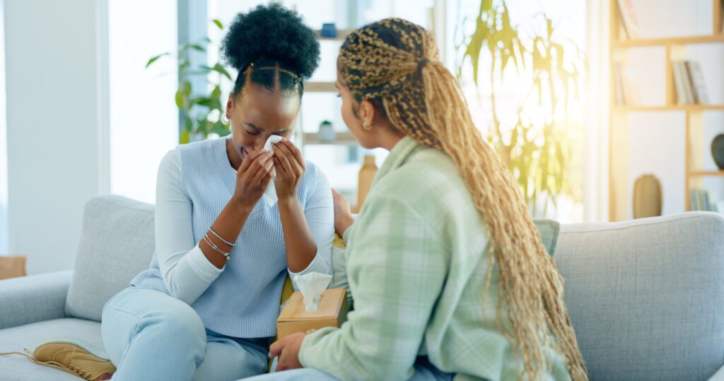 Sad, empathy and support with black woman friends on a sofa in the living room of a home together. Depression, mental health and a young person crying into a tissue during loss, pain or grief